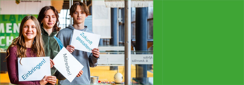 Drei Jugendliche stehen im Bürgerfoyer des Landtags mit Schildern, die dazu auffordern, aktiv zu werden.